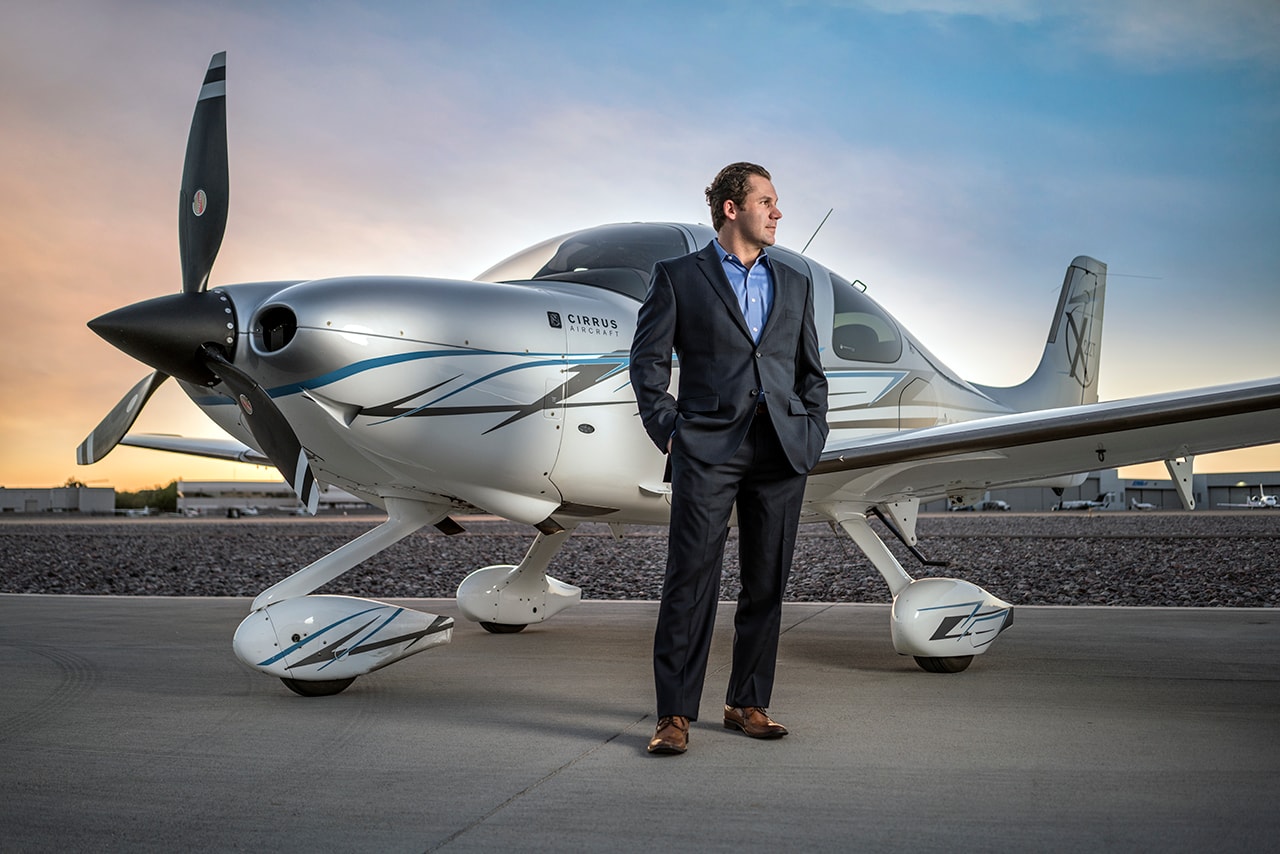 Man in suit standing before Cirrus aircraft on tarmac at sunset