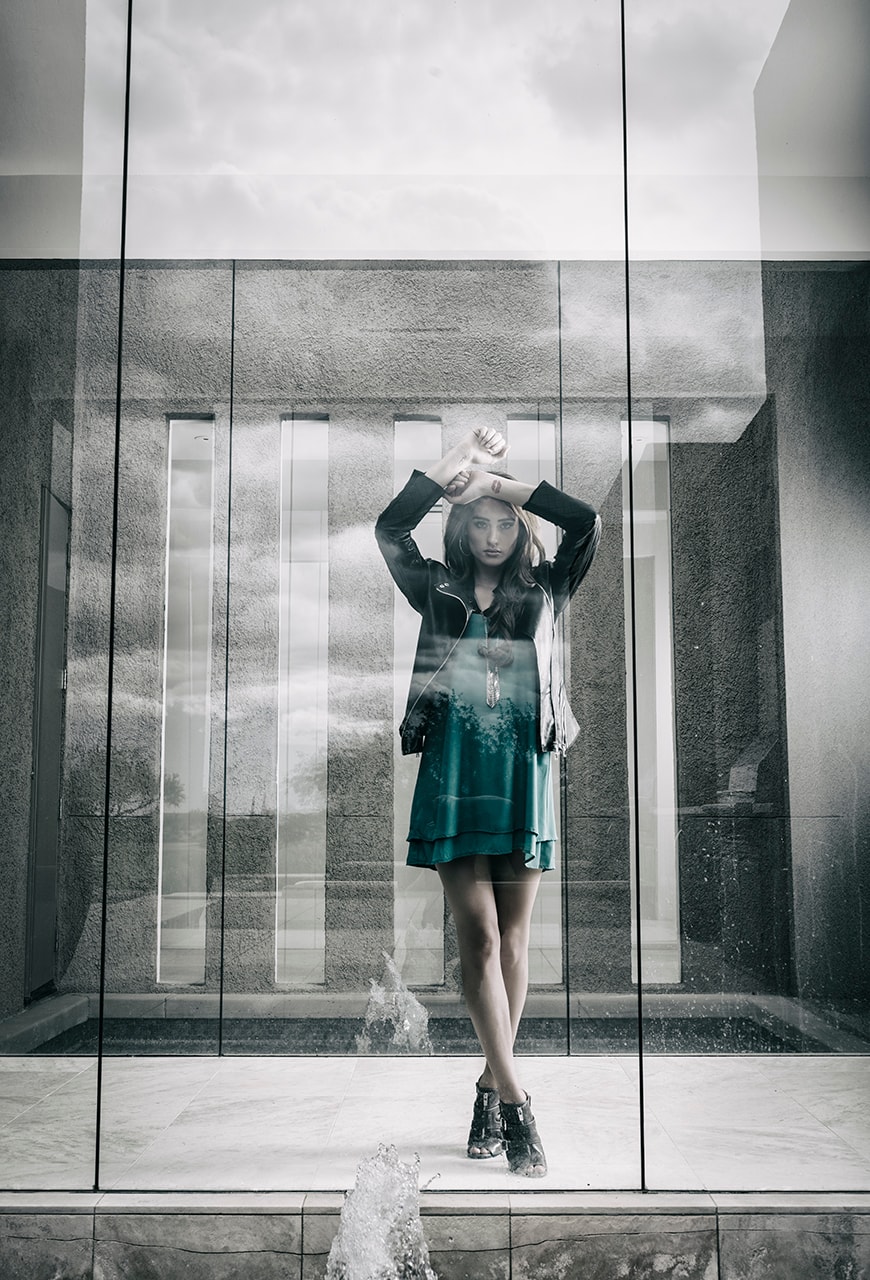 Woman in teal dress posing in front of glass architectural facade with water splash