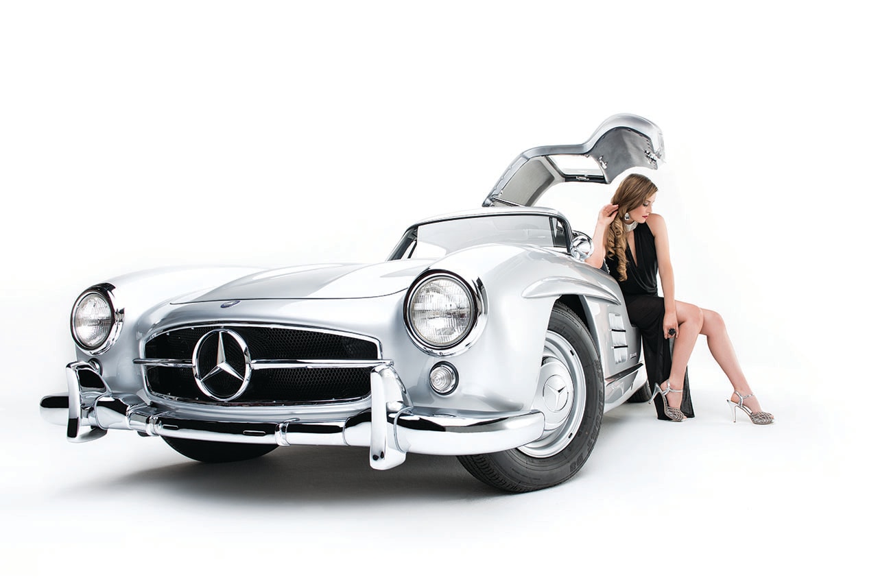 Woman in black dress seated beside silver Mercedes 300SL gullwing on white background