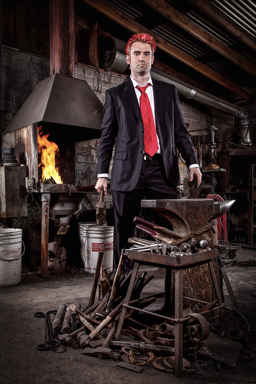 Man with red hair in suit standing at forge in blacksmith workshop